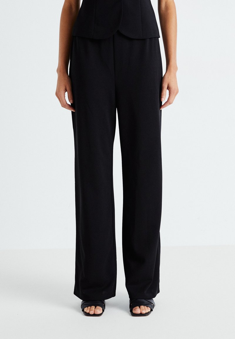 Pantalons larges noirs en tissu lisse, présentant une coupe décontractée et une taille élastique. Associés à des sandales noires simples.
