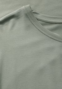 Ljust grönt bomullstyg med en mjuk textur, med rund halsringning och subtila sömnadsdetaljer längs kanterna.