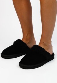Zwarte instapslippers met een zachte, gestructureerde zwarte bontvoering en een platte rubberen zool, ontworpen voor comfort en gemak.