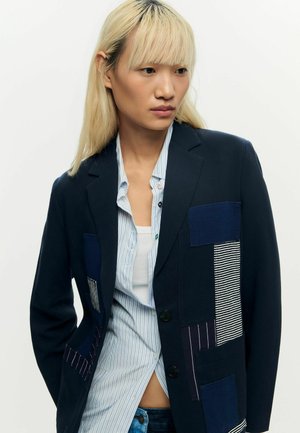 Jonge persoon met lang blond haar, gekleed in een marineblauw blazer met patchworkdetails over een gestreept shirt en een wit hemdje.