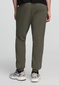 Oliwkowe dresy z miękkiego materiału o zwężającym się kroju, z elastycznym pasem i bocznymi kieszeniami, połączone z szarymi trampkami.