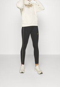 Krämfärgad hoodie med en texturerad logotyp, tillsammans med figurnära svarta leggings med guldstripes på sidorna och varumärkesdetaljer. Avslutad med lätta sneakers.