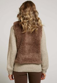 Femme aux cheveux ondulés portant un pull beige et un gilet en fourrure synthétique marron, vue de dos sur un fond uni.