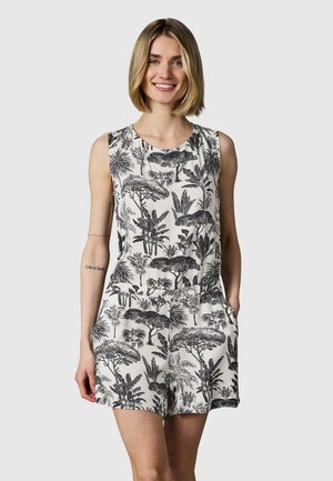 Smilende kvinde med blond bob iført hvid ærmeløs romper med sort tropisk træ- og planteprint, hænder afslappede, lysegrå baggrund.