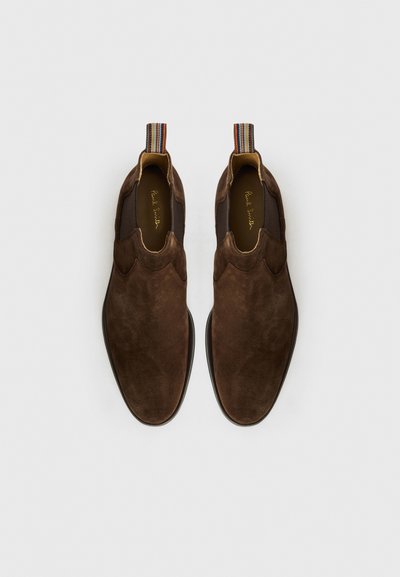 Zapatos sin cordones de ante marrón que presentan un diseño ondulado, una textura suave y una asa a rayas en el talón.