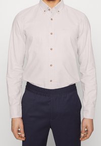 Camicia button-down beige chiaro con bottoni marroni e un logo discreto, abbinata a pantaloni blu navy con vita elastica. Tessuto di cotone liscio.