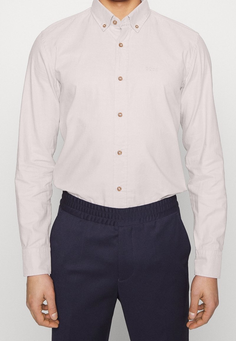 Camicia button-down beige chiaro con bottoni marroni e un logo discreto, abbinata a pantaloni blu navy con vita elastica. Tessuto di cotone liscio.