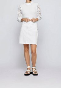 Kvinna iklädd en vit stickad topp med ögla spetsärm, vit knälång kjol och vita sandaler med svarta sulor och ankelremmar.