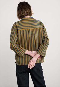 Camisa a rayas en azul y mostaza con un pliegue en la espalda, mangas tres cuartos y cuello, combinada con pantalones negros.