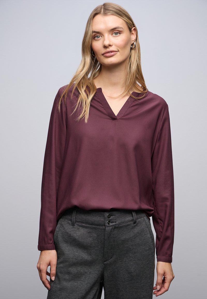 Bordeauxfarbene Bluse mit langen Ärmeln und V-Ausschnitt, aus glattem Material. Kombiniert mit dunkelgrauen, taillierten Hosen mit zwei vorderen Knöpfen.