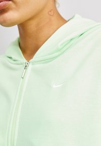 Mátově zelená zipová mikina vyrobená z měkké látky, s bílým logem Nike a jemným tetováním srdce na krku.