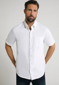 Camisa blanca de manga corta, con botones y un bolsillo, que presenta un cuello y una textura sutil, combinada con jeans oscuros.