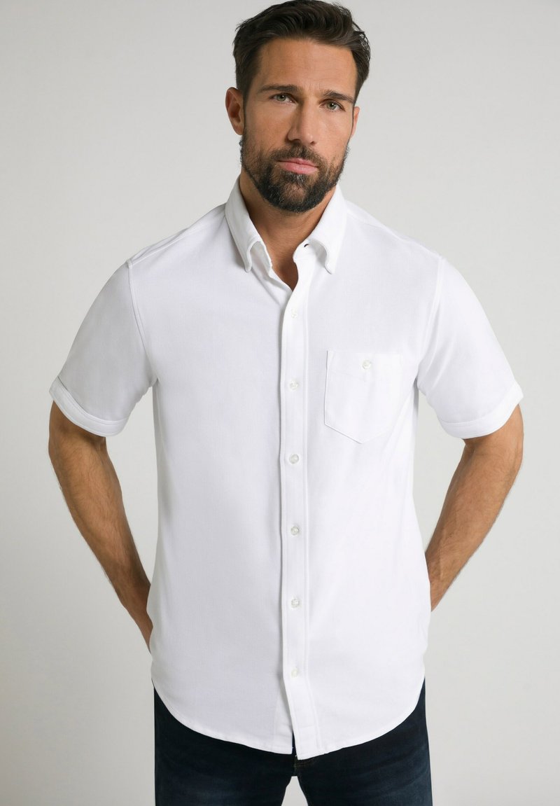Camisa blanca de manga corta, con botones y un bolsillo, que presenta un cuello y una textura sutil, combinada con jeans oscuros.
