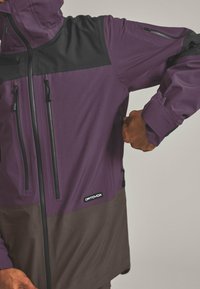 Purple and black waterproof jacket with zippered pockets, adjustable cuffs, and a short hem. Features a logo on the lower front.