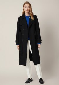 Czarny podwójnie zapinany płaszcz z wyłogowym kołnierzem, długimi rękawami i paskiem, nałożony na niebieski sweter, w połączeniu z białymi spodniami i mokasynami.