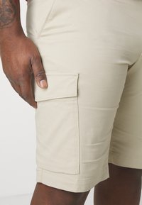 Pantaloni cargo beige realizzati in tessuto liscio, con tasca laterale e risvolti. Design semplice e funzionale con minimi dettagli di cucitura.