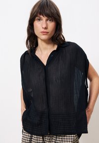 Schwarze, halbtransparente Bluse mit Knopfleiste aus strukturiertem Stoff und kragenlosem Ausschnitt. Weite Ärmel und lockere Passform mit einem dezenten Kräuselmuster.