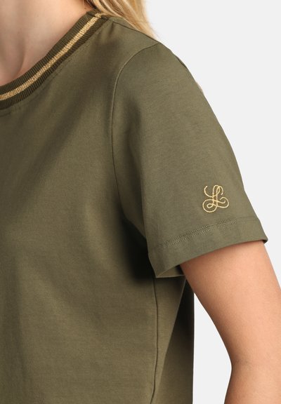 Femme portant une chemise à manches courtes vert olive avec une bordure dorée sur le col et un logo brodé en or sur la manche, sur fond uni.