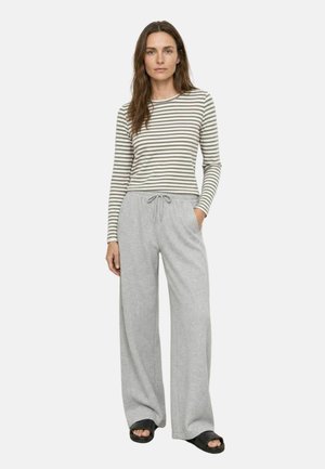 Vrouw die een grijs-wit gestreept shirt met lange mouwen draagt, lichtgrijze wijde broek met trekkoord en zwarte sandalen met open tenen.