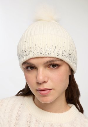 Giovane donna che indossa un berretto di maglia color crema con pompon decorato con piccole strass argentate e un maglione di maglia a trecce color crema.