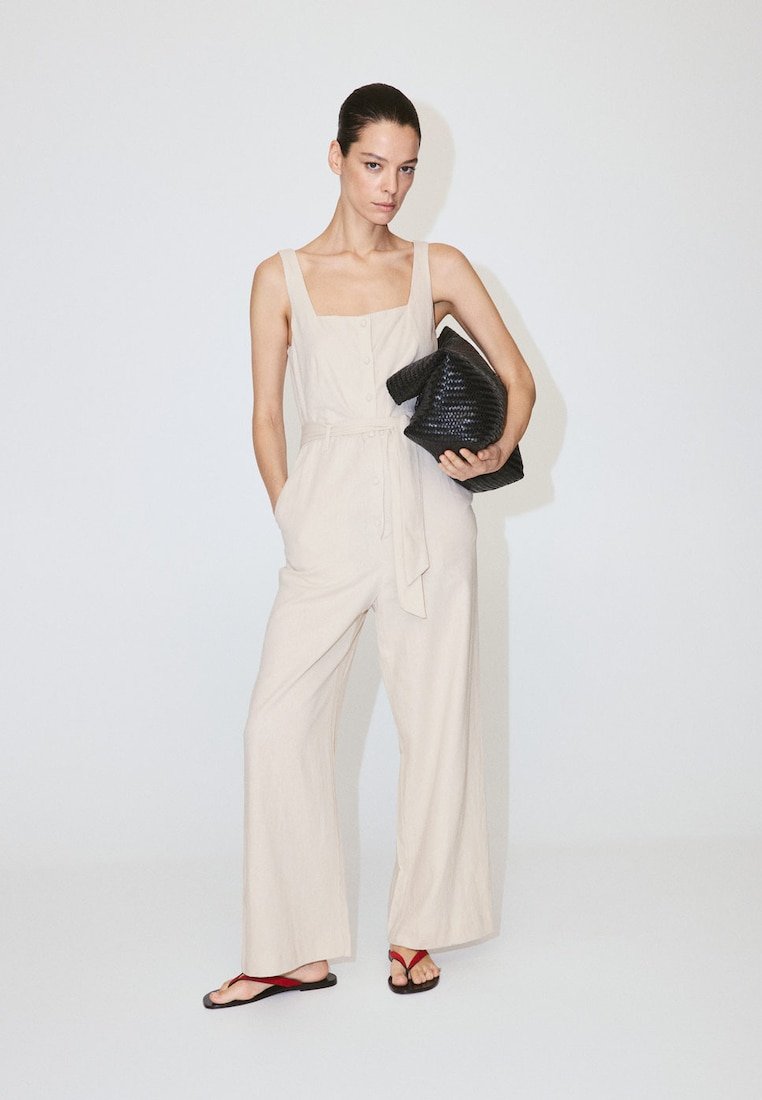Femme portant une combinaison beige sans manches et des sandales rouges, tenant une grande pochette noire tissée sur fond blanc uni.