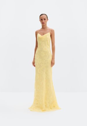 Femeie purtând o rochie lungă din dantelă florală de culoare galben pal, cu bretele subțiri, stând în picioare pe un fundal alb simplu.