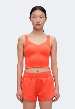 Jeune femme portant un crop top orange vif et un short de sport assorti, face à un fond clair uni.