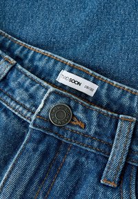 Denim džinsi vidēji zilā krāsā ar redzamām šuvēm, zīmolu metāla pogu aizdari un birku ar uzrakstu "TWO SOON 28/32."