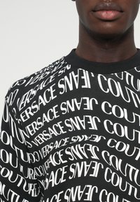Črna kapuca s ponavljajočim se belim besedilnim vzorcem "Versace Jeans Couture" v različnih orientacijah, z rebrastim krožnim ovratnikom.