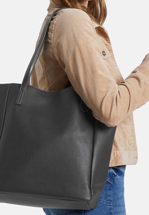 Svart texturerad läderhandväska med två långa handtag, slapp design och stabil botten, hålls av en person som bär en manchesterjacka.