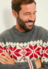 Mann mit Bart, der lächelt und nach unten schaut, trägt einen grauen Strickpullover mit roten und weißen Schneeflockenmuster, die Arme verschränkt.