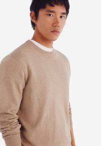 Maglione beige a girocollo in un morbido tessuto a maglia; polsini e orlo a costine; design aderente con leggero stretch; indossato sopra una camicia bianca.
