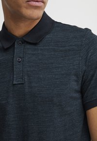 Chemise polo bleu foncé en tissu texturé. Présente un col noir, une patte de boutonnage à trois boutons et des manches courtes avec des poignets côtelés.