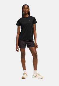 T-shirt noire à col rond fabriquée en tissu léger, assortie à un short sombre. Le modèle porte des chaussures de sport multicolores avec des semelles texturées.