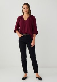 Frau steht mit der Hand in der Tasche, trägt eine burgunderrote Bluse mit langen Ärmeln, schwarze Hosen mit Gürtel, schwarze Slipper und eine goldene Halskette.