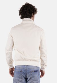 Giacca leggera in crema con colletto alto, polsini e vita elastici. Presenta una texture liscia e senza hardware o motivi visibili.