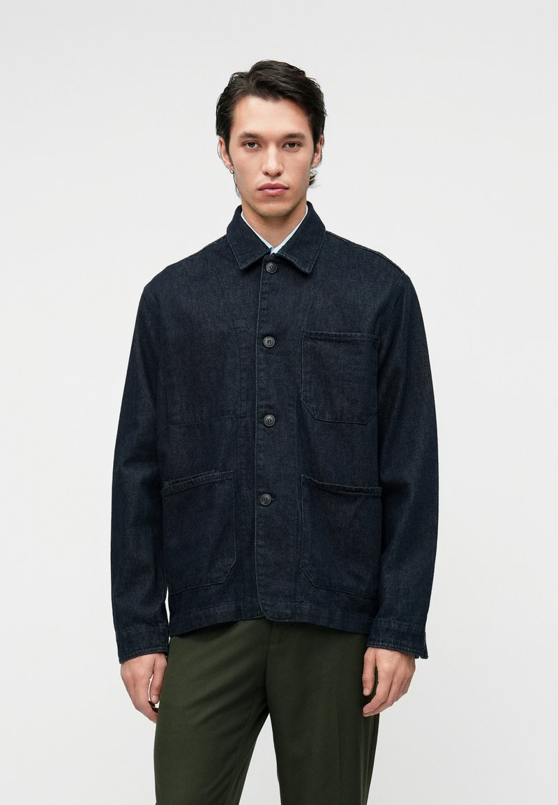 Tamsiai mėlynas džinsinis švarkas su apykakle, priekyje su mygtukais ir dviem krūtinės kišenėmis. Turi laisvą fasoną ir tekstūruotą medžiagą.