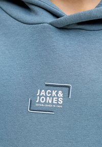 Κοντινό πλάνο στο στήθος ενός μπλε φούτερ με λευκή επιγραφή "JACK & JONES ESTABLISHED IN 1990" και καμπούρα κουκούλας διπλωμένη.