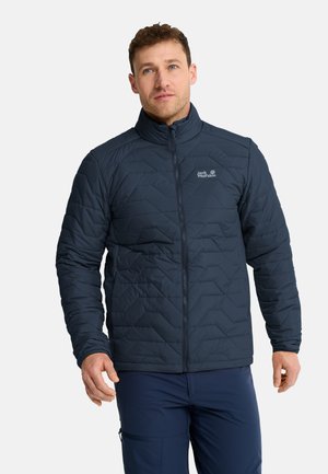 Mann trägt eine dunkelblaue gesteppte Jacke mit Reißverschluss und hohem Kragen sowie dunkelblaue Hose, steht vor einem schlichten hellen Hintergrund.