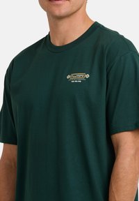 Hombre vistiendo una camiseta de manga corta verde oscuro con un pequeño logo blanco y amarillo de Billabong en el lado izquierdo del pecho.