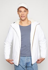 Giacca bianca con zip e foderata in pile, con polsini elastici e zip di accento gialla. Indossata su una maglietta grigia e jeans in denim.