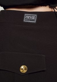 Vêtement noir avec un patch Versace Jeans Couture et une poche avec un bouton doré gravé du nom de la marque.