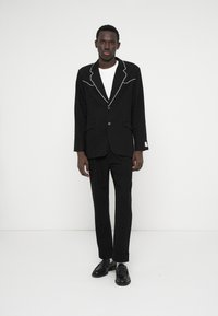 Czarny garnitur w kropki z białym wykończeniem, z pojedynczym zapięciem na guzik, dopasowany krój, w połączeniu z czarnymi mokasynami.