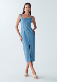 Zila midi kleita bez piedurknēm ar kvadrātveida izgriezumu un priekšpuses šķēlumu. Vidū ir pogas un gluda tekstūra. Kopa ar pītām sandales.