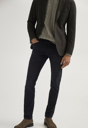 Homme portant un blazer gris foncé, un pull en maille torsadée gris clair, un pantalon slim foncé et des bottes en daim marron, debout avec une main dans la poche.
