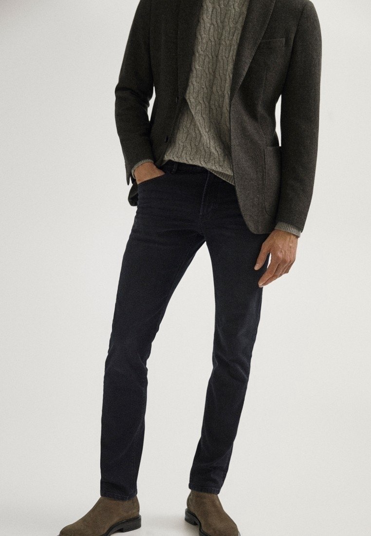 Homme portant un blazer gris foncé, un pull en maille torsadée gris clair, un pantalon slim foncé et des bottes en daim marron, debout avec une main dans la poche.