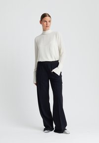 Witte blouse met lange mouwen en hoge hals; gecombineerd met zwarte wijde broek. Visuele textuur contrasteert met strakke lijnen en vloeiende stoffen.