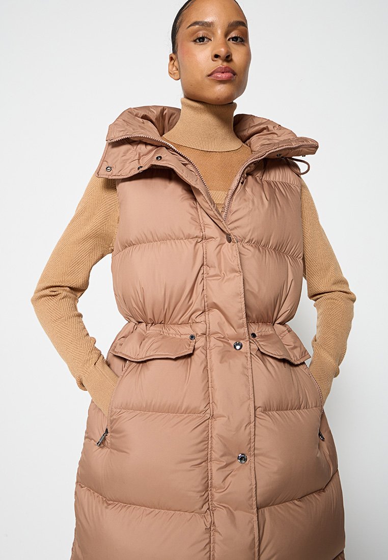 Langer, brauner Daunenweste mit Kapuze, ausgestattet mit einem Gürtel in der Taille, Seitentaschen und einem strukturierten beigen Pullover darunter. Glatte, glänzende Oberfläche.