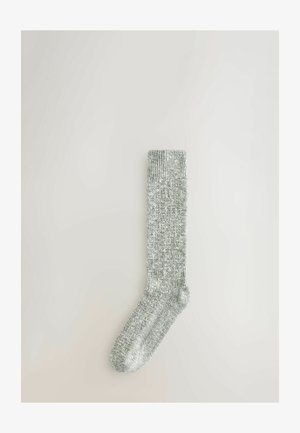 Yksi harmaa neulottu sukka, jossa on ribbireuna, aseteltuna litteäksi vaaleaa neutraalia taustaa vasten.