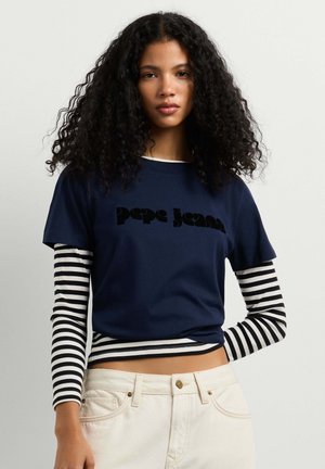 Donna con capelli ricci che indossa una t-shirt Pepe Jeans blu navy sovrapposta a una maglia a maniche lunghe a righe bianche e nere e pantaloni beige chiaro.